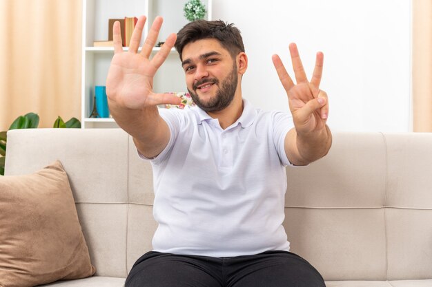 Giovane uomo in abiti casual che sembra felice e sorridente positivo che mostra il numero otto seduto su un divano in un soggiorno luminoso