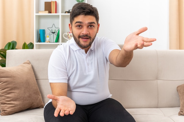 Giovane uomo in abiti casual che sembra felice ed eccitato con le braccia aperte seduto su un divano in un soggiorno luminoso