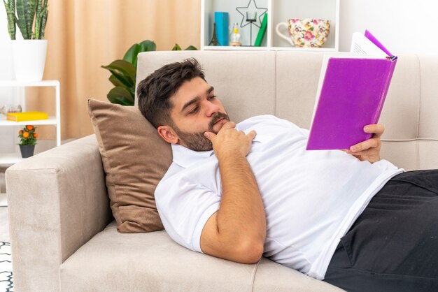 Giovane uomo in abiti casual che tiene in mano un libro che legge con una faccia seria che trascorre il fine settimana a casa sdraiato su un divano in un soggiorno luminoso