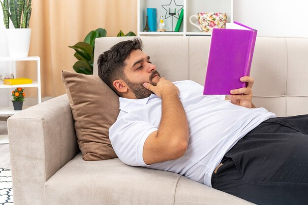 Giovane uomo in abiti casual che tiene in mano un libro che legge con una faccia seria che trascorre il fine settimana a casa sdraiato su un divano in un soggiorno luminoso