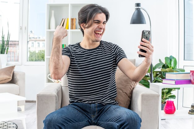 Giovane uomo in abiti casual che tiene smartphone guardandolo felice ed eccitato alzando il pugno seduto sulla sedia in un soggiorno luminoso