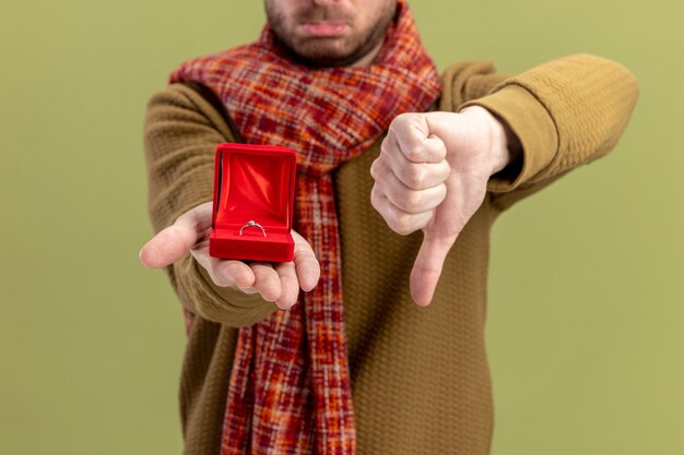 Giovane uomo in abiti casual con sciarpa intorno al collo che mostra un riquadro rosso con anello di fidanzamento che mostra i pollici verso il basso il giorno di San Valentino concetto in piedi su sfondo verde