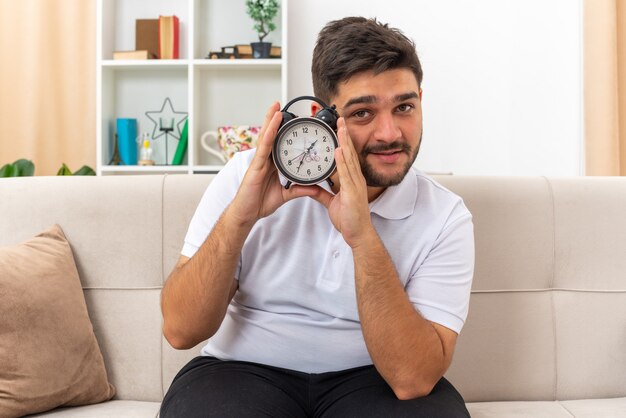 Giovane uomo in abiti casual con sveglia guardando con un sorriso sul viso felice e positivo seduto su un divano in un soggiorno luminoso