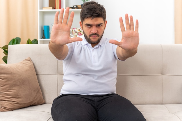 Giovane uomo in abiti casual con una faccia seria che fa un gesto di arresto con le mani seduto su un divano in un soggiorno luminoso