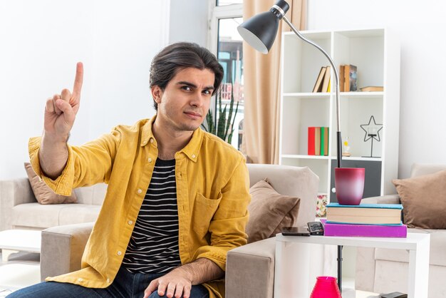 Giovane uomo in abiti casual felice e fiducioso che mostra il dito indice che ha una buona idea seduto sulla sedia in un soggiorno luminoso