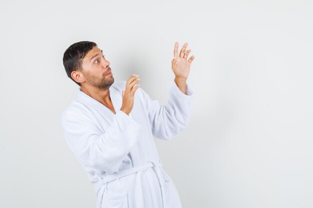 Giovane uomo in accappatoio bianco alzando le mani in modo protettivo e guardando spaventato, vista frontale.