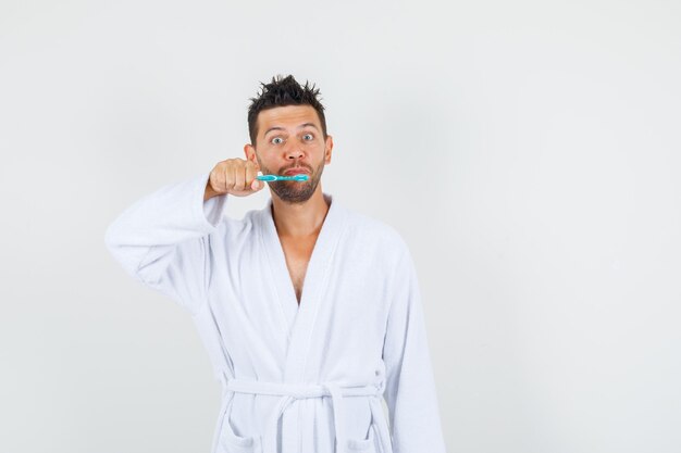 Giovane uomo in accappatoio bianco tenendo uno spazzolino da denti, vista frontale.