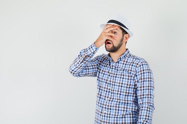 Giovane uomo in camicia a quadri, cappello guardando attraverso le dita e guardando sorpreso, vista frontale.