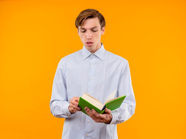 Giovane uomo in camicia bianca che tiene libro aperto guardandolo con faccia seria in piedi sopra la parete arancione