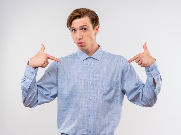 Giovane uomo in camicia blu che punta a se stesso chiedendo in piedi sul muro bianco