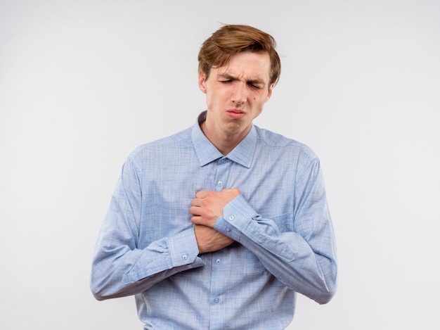 Giovane uomo in camicia blu che tiene hnads sul petto alla ricerca di malessere sensazione di dolore in piedi sopra il muro bianco