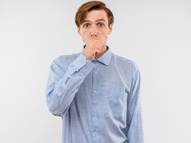 Giovane uomo in camicia blu confuso che copre la bocca con il pugno in piedi sul muro bianco