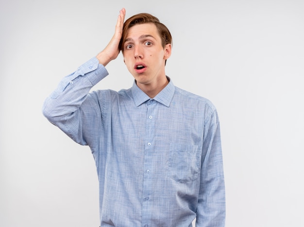 Giovane uomo in camicia blu confuso e preoccupato con la mano sulla sua testa per errore in piedi sopra il muro bianco