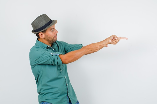 Giovane uomo in camicia, cappello, jeans che punta il dito lontano e che guarda concentrato