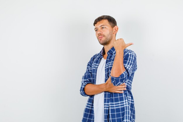 Giovane uomo in camicia che punta indietro con il pollice e sorridente, vista frontale.