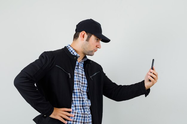 Giovane uomo in camicia, giacca, berretto parlando in chat video e guardando serio