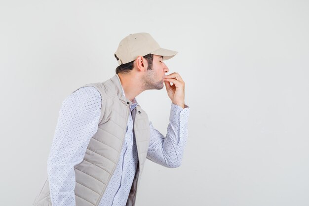 Giovane uomo in camicia, giacca senza maniche, berretto che mostra un gesto delizioso e che sembra concentrato, vista frontale.