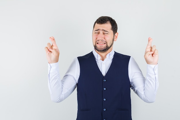 Giovane uomo in camicia, giubbotto tenendo le dita incrociate e guardando speranzoso