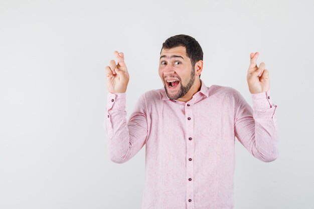 Giovane uomo in camicia rosa tenendo le dita incrociate e guardando allegro