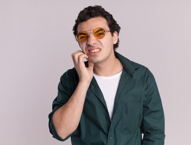 Giovane uomo in camicia verde con gli occhiali cercando confuso grattandosi la faccia in piedi sul muro bianco