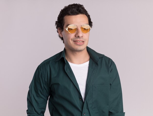Giovane uomo in camicia verde con gli occhiali che guarda da parte con il sorriso sul viso in piedi sul muro bianco