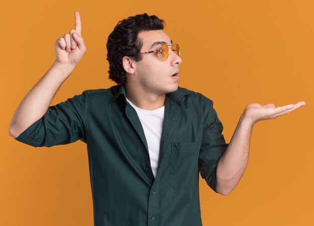 Giovane uomo in camicia verde con gli occhiali che guarda da parte sorpreso indicando con il dito indice in su che presenta con lo spazio della copia del braccio che sta sopra la parete arancione