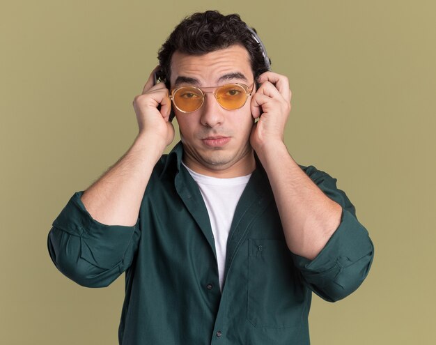 Giovane uomo in camicia verde con gli occhiali con le cuffie guardando davanti confuso in piedi sopra la parete verde