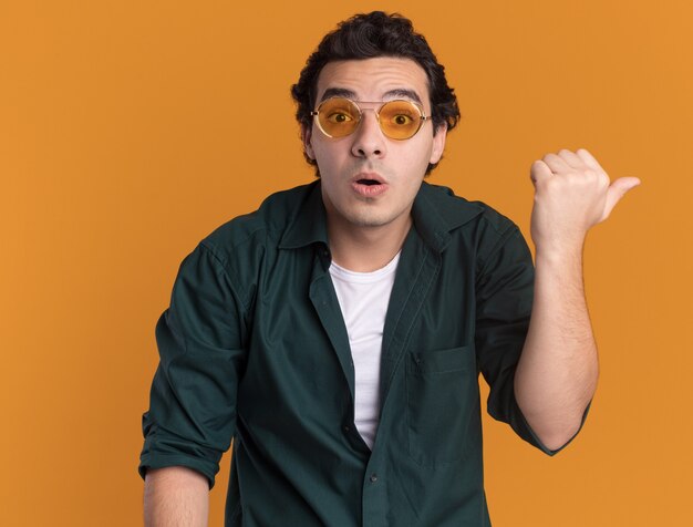 Giovane uomo in camicia verde con gli occhiali guardando davanti confuso che punta con il pollice a lato in piedi sopra la parete arancione