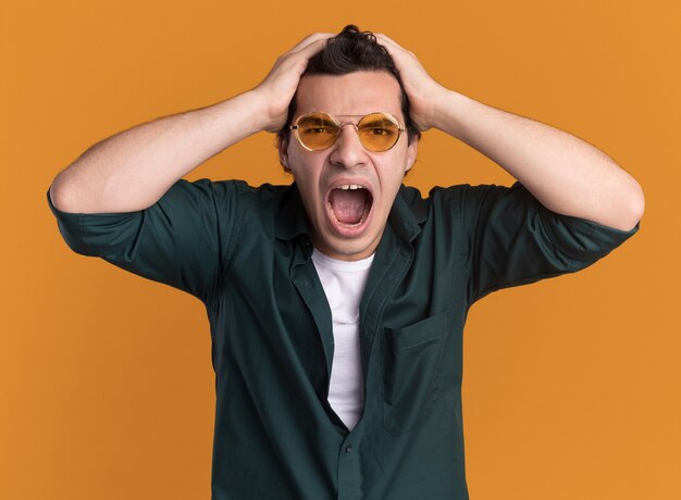 Giovane uomo in camicia verde con gli occhiali guardando davanti pazzo pazzo e arrabbiato tirando i capelli in piedi sopra la parete arancione