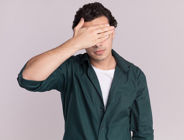 Giovane uomo in camicia verde stanco e annoiato chiudendo gli occhi con la mano in piedi sul muro bianco