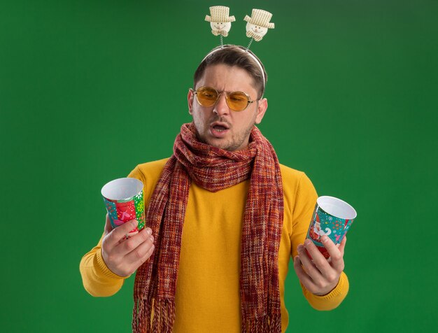 Giovane uomo in dolcevita giallo con sciarpa calda e occhiali che indossa un bordo divertente sulla testa che tiene due tazze colorate cercando confuso avendo dubbi in piedi sul muro verde