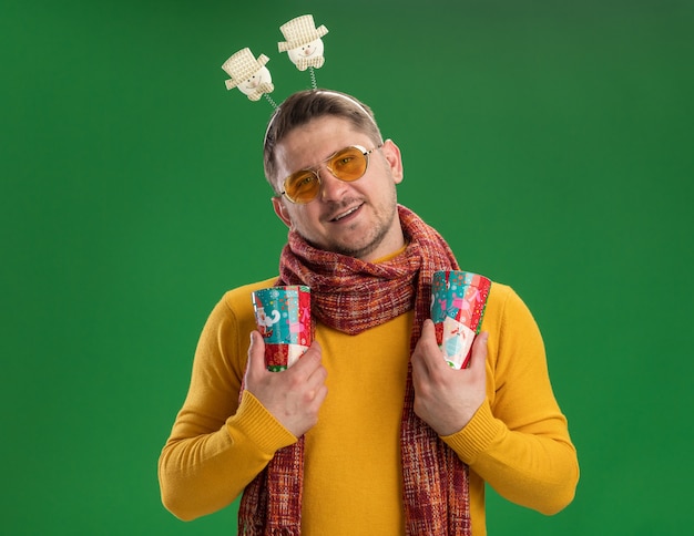 Giovane uomo in dolcevita giallo con sciarpa calda e occhiali indossando il bordo divertente sulla testa tenendo tazze colorate con il sorriso sul viso in piedi sopra la parete verde