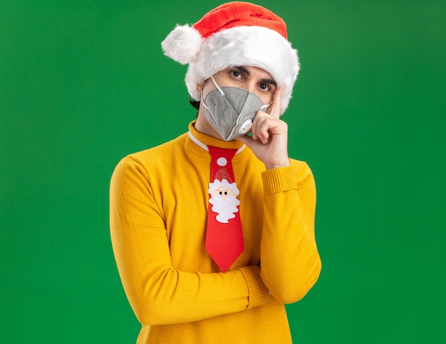 Giovane uomo in dolcevita giallo e cappello da Babbo Natale con cravatta divertente che indossa la maschera protettiva per il viso che guarda l'obbiettivo con la faccia seria con la mano sul mento pensando in piedi su sfondo verde