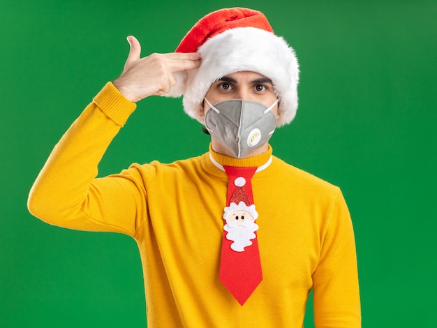 Giovane uomo in dolcevita giallo e cappello da Babbo Natale con cravatta divertente che indossa una maschera protettiva facciale che fa il gesto della pistola sul tempio in piedi su sfondo verde green