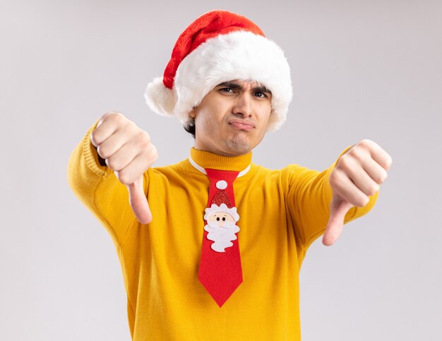 Giovane uomo in dolcevita giallo e cappello da Babbo Natale con cravatta divertente guardando la telecamera scontento che mostra i pollici verso il basso in piedi su sfondo bianco