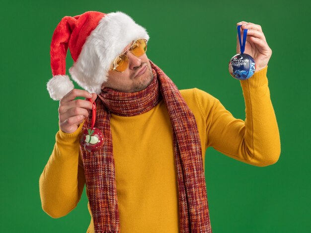 Giovane uomo in dolcevita giallo e cappello da Babbo Natale con gli occhiali che tengono i giocattoli per l'albero di natale guardandoli con espressione confusa avendo dubbi in piedi sopra la parete verde