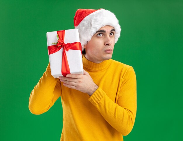 Giovane uomo in dolcevita giallo e cappello da Babbo Natale in possesso di un regalo sopra l'orecchio cercando intrigato in piedi su sfondo verde