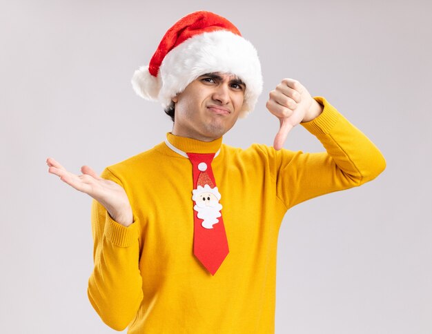 Giovane uomo in dolcevita giallo e cappello della santa con cravatta divertente guardando la telecamera che presenta qualcosa con il braccio della sua mano che mostra i pollici verso il basso essendo dispiaciuto in piedi su sfondo bianco