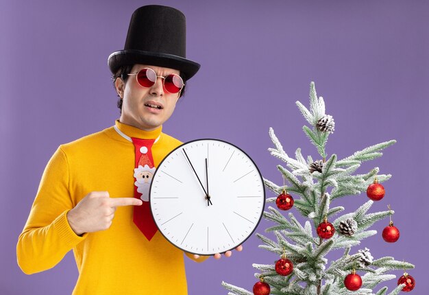 Giovane uomo in dolcevita giallo e occhiali che indossa cappello nero tenendo l'orologio da parete che punta con il dito indice contro di esso con la faccia seria in piedi accanto a un albero di natale oltre il muro viola