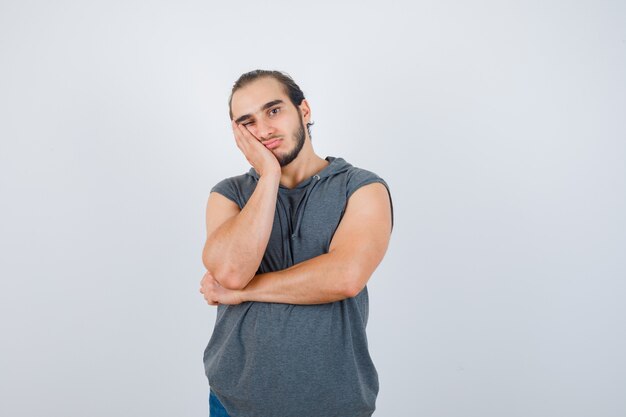 Giovane uomo in felpa con cappuccio senza maniche tenendo la mano sulla guancia e guardando pensieroso, vista frontale.