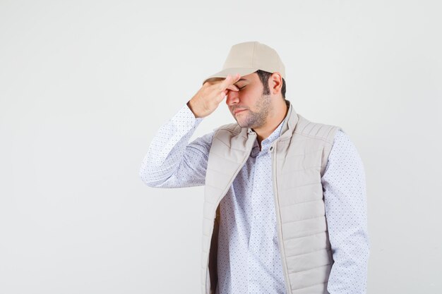 Giovane uomo in giacca beige e berretto che copre parte del viso con la mano e sembra stanco, vista frontale.