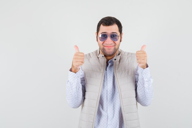 Giovane uomo in giacca beige e berretto indossare occhiali e mostrando il doppio pollice in alto e guardando ottimista, vista frontale.