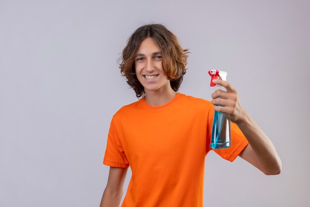 Giovane uomo in maglietta arancione azienda spray per la pulizia guardando la fotocamera sorridendo allegramente felice e positivo in piedi su sfondo bianco