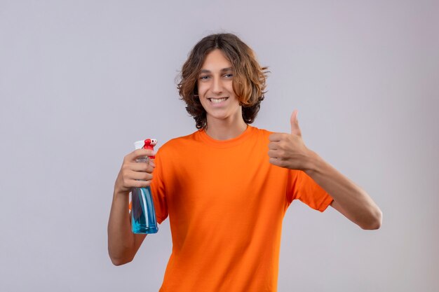 Giovane uomo in maglietta arancione azienda spray per la pulizia sorridendo allegramente guardando la telecamera che mostra i pollici in su in piedi su sfondo bianco