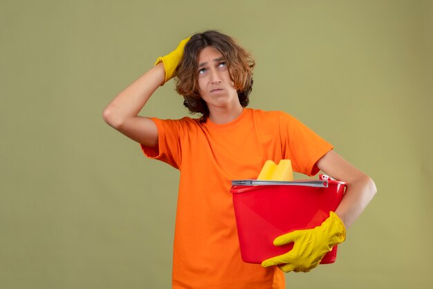 Giovane uomo in maglietta arancione che indossa guanti di gomma tenendo la benna con strumenti di pulizia guardando in alto con la mano sulla testa per errore cercando di pensare confuso in piedi su sfondo verde