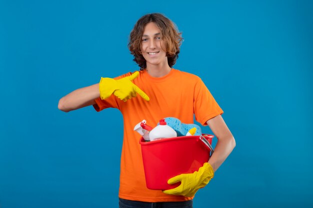 Giovane uomo in maglietta arancione che indossa guanti di gomma tenendo la benna con strumenti di pulizia puntando con il dito ad esso sorridendo allegramente felice e positivo in piedi su sfondo blu