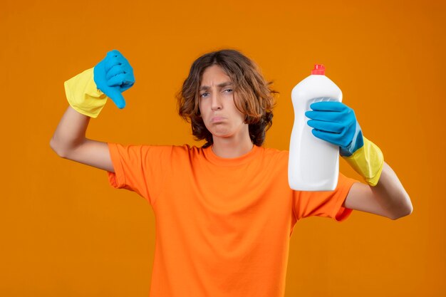 Giovane uomo in maglietta arancione che indossa guanti di gomma tenendo la bottiglia con prodotti per la pulizia che mostra i pollici verso il basso con l'espressione triste sul viso in piedi su sfondo giallo
