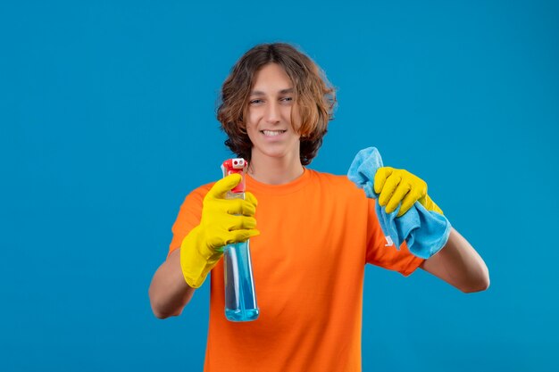 Giovane uomo in maglietta arancione che indossa guanti di gomma tenendo spray per la pulizia e tappeto guardando la fotocamera con un sorriso fiducioso in piedi su sfondo blu