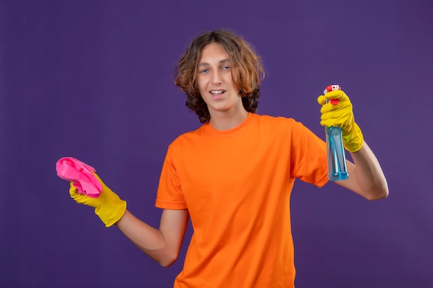 Giovane uomo in maglietta arancione che indossa guanti di gomma tenendo spray per la pulizia e tappeto guardando la fotocamera con un sorriso fiducioso in piedi su sfondo viola