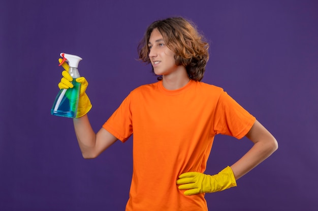 Giovane uomo in maglietta arancione che indossa guanti di gomma tenendo spray per la pulizia guardando da parte sorridente con la faccia felice pronta per pulire in piedi su sfondo viola